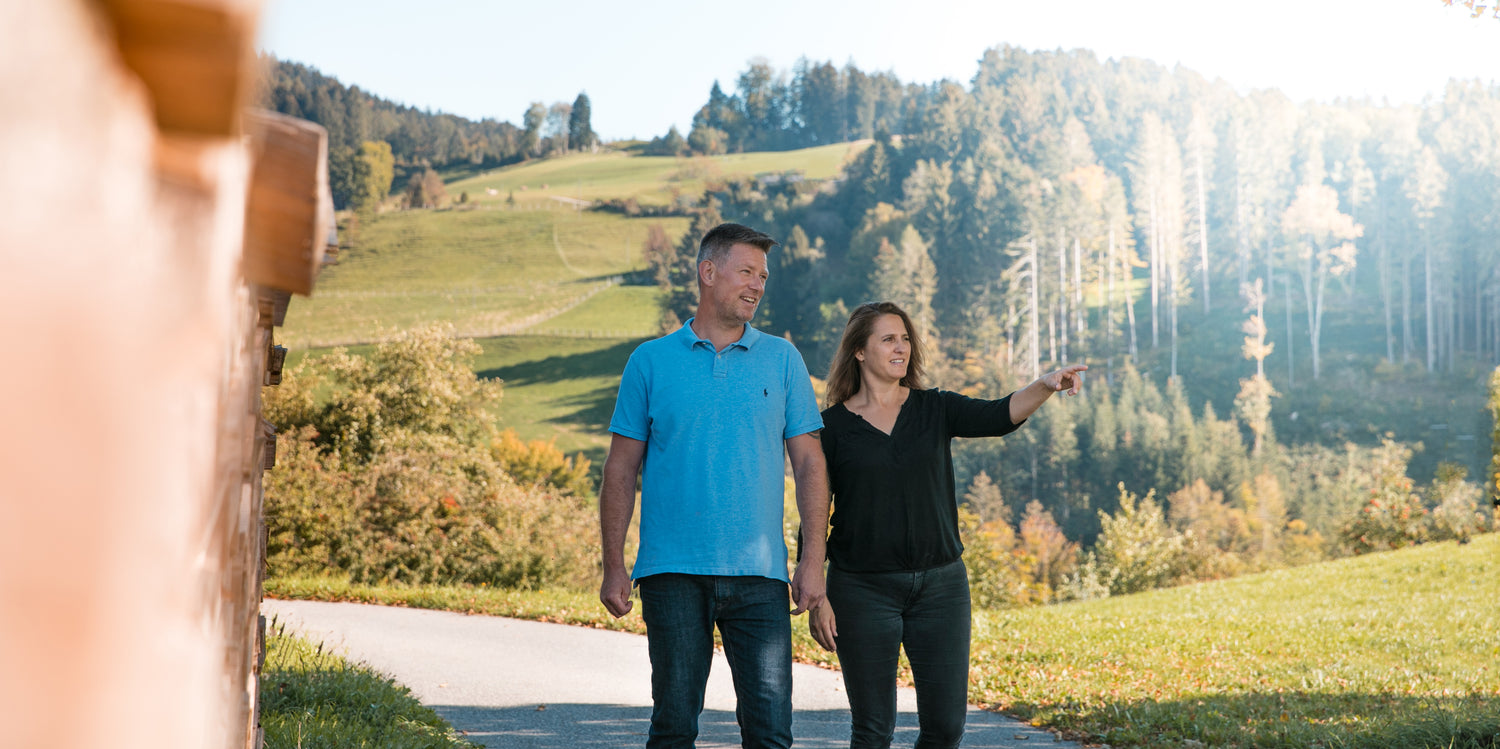 Couple walking outdoors in casual blue polo and black top, surrounded by green hills and trees in autumn colors.