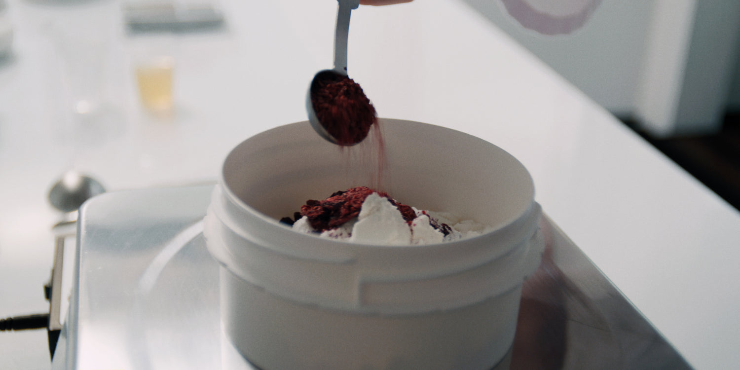 Scoop of vibrant red protein powder being added to a white container with a white powder blend on a scale.