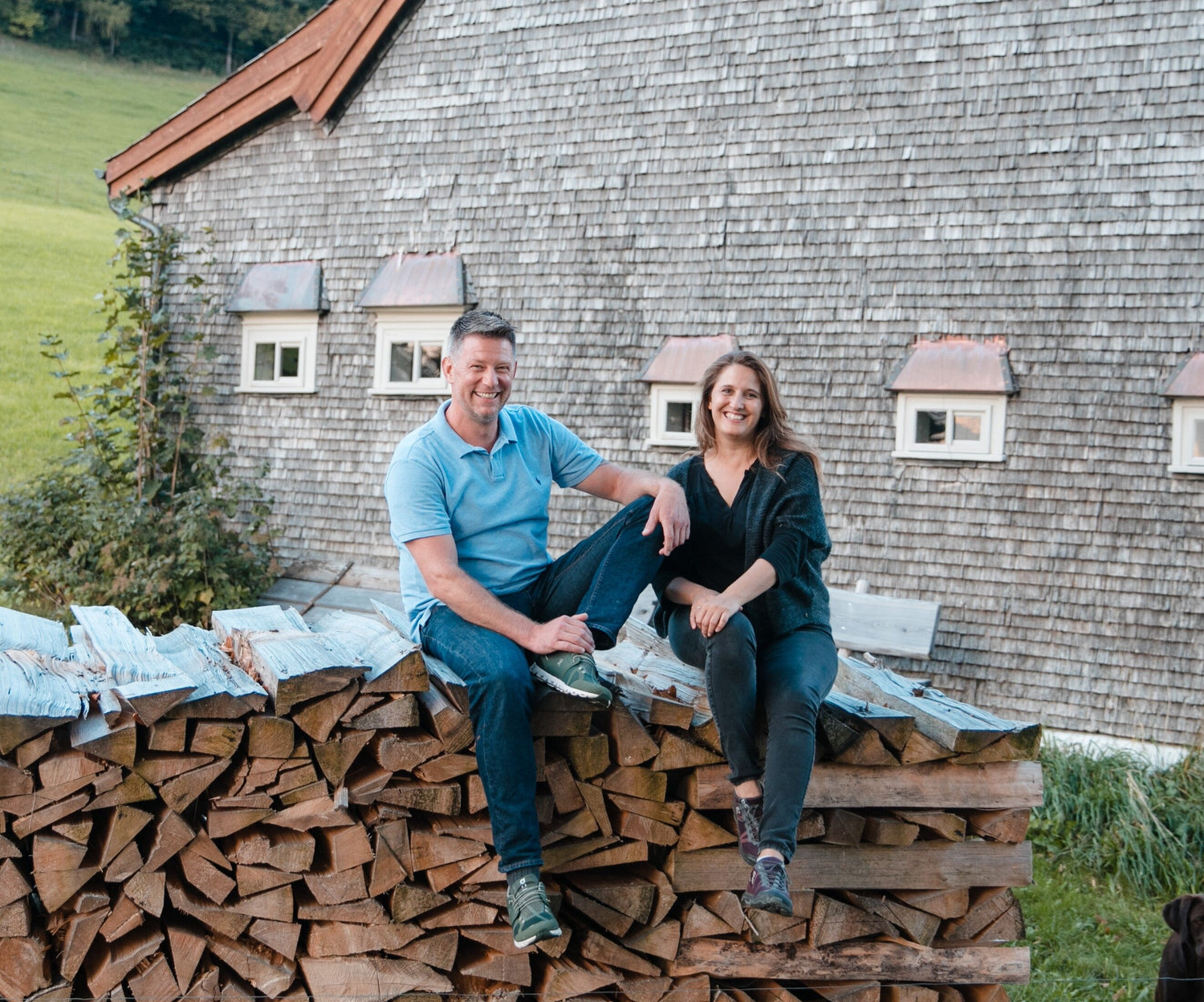 Couple sitting on stacked wood, wearing casual blue shirt and black sweater, in front of rustic wooden barn.